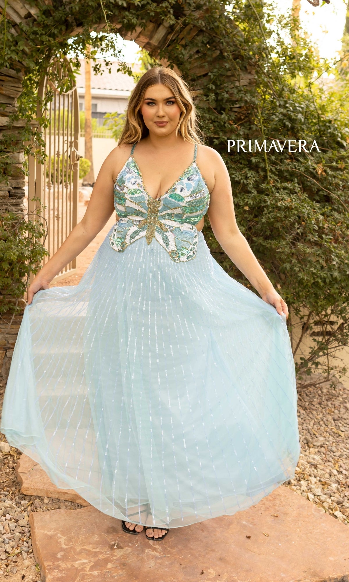 A woman in the Plus Size Butterfly Prom Dress 14011 by Primavera poses outdoors under a stone archway with greenery and